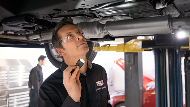 Mechanic inspects car undercarriage with flashlight in auto service garage, wearing "Certified Service" uniform.