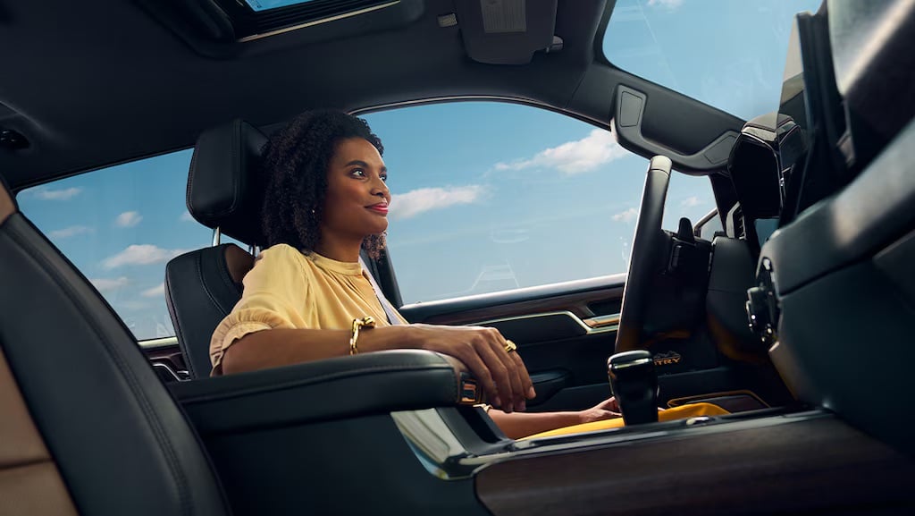 Woman in a beige blouse driving a car, looking content. Blue sky visible through the window.