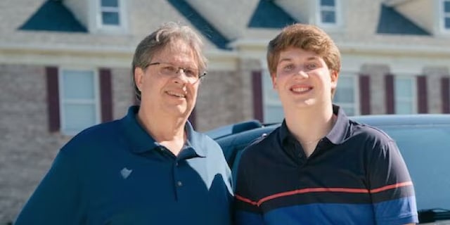 Two people smiling in front of a suburban house on a sunny day.