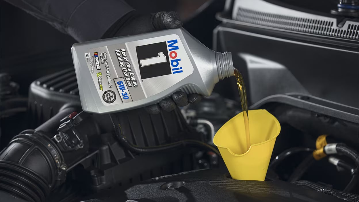 Pouring Mobil 1 motor oil into engine with yellow funnel in car maintenance scene.