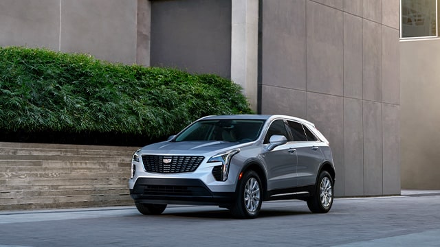 Silver Cadillac SUV parked next to modern building facade and lush greenery.