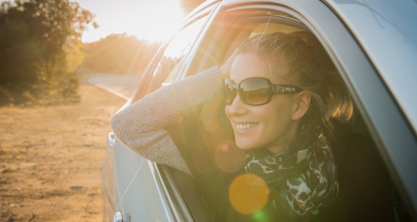 Woman smiling, wearing sunglasses, leans out car window enjoying sunlight on a rural road.