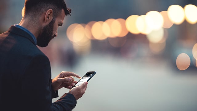 Man using smartphone outdoors, evening city lights blurred in background.