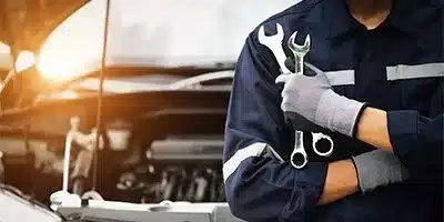 Mechanic holding wrenches in front of an open car hood, wearing navy uniform and gloves.
