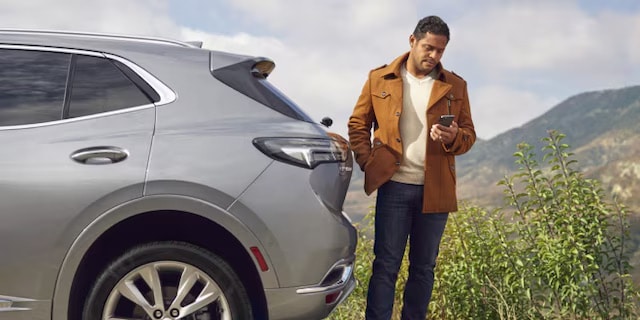 Man in a brown coat checks his phone beside a silver SUV, with mountains in the background.
