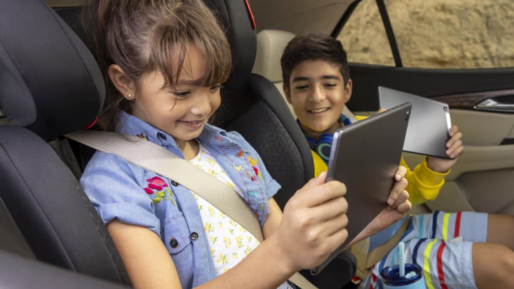 Children enjoying tablets in a car, smiling, wearing seatbelts and colorful clothing.
