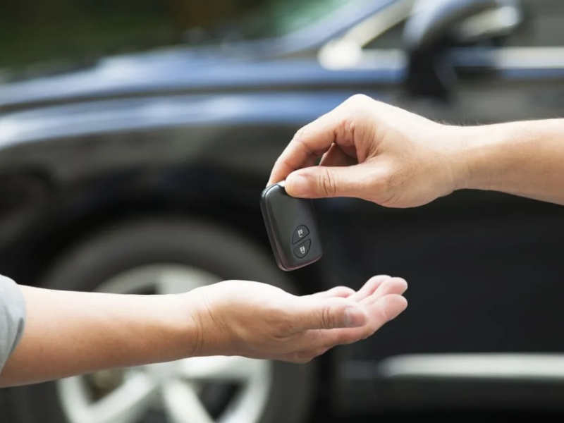 Hand giving a car key fob to another person with a blurred vehicle in the background.